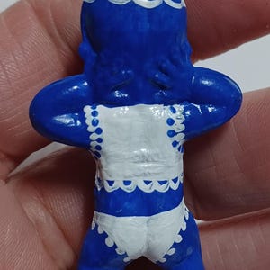 May include: A blue figurine of a baby with white accents. The figurine has a white bib and diaper with scalloped edges. The baby is holding its arms up to its head. A silver loop is at the top for hanging.