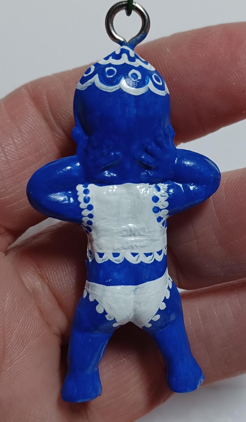 May include: A blue figurine of a baby with white accents. The figurine has a white bib and diaper with scalloped edges. The baby is holding its arms up to its head. A silver loop is at the top for hanging.