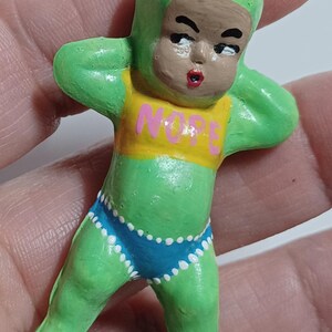May include: A green figurine of a baby-like figure with a green cap and body suit, brown face, and red lips. The figure wears a yellow top with the word "NOPE" and blue shorts with white polka dots. A silver loop is attached to the top.