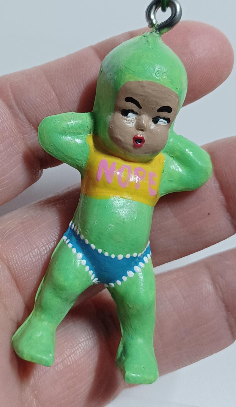 May include: A green figurine of a baby-like figure with a green cap and body suit, brown face, and red lips. The figure wears a yellow top with the word "NOPE" and blue shorts with white polka dots. A silver loop is attached to the top.