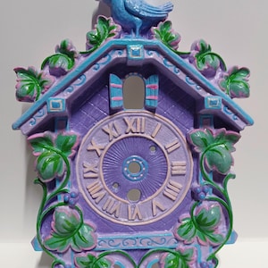 May include: A decorative cuckoo clock in shades of purple, blue, and green. The clock features Roman numerals, a bird on top, and floral accents. The clock is a whimsical, decorative item.