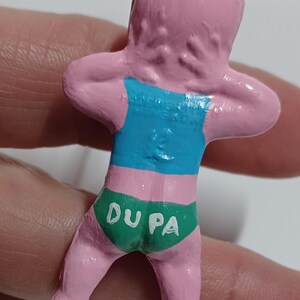 May include: A pink figurine of a person with arms raised, wearing a blue top and green underwear. The underwear has the text "DU PA" in white. The figurine is attached to a silver-colored loop.