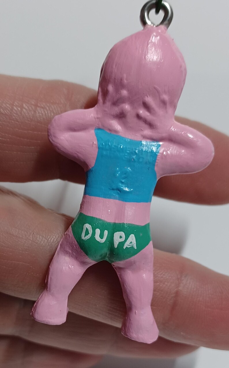 May include: A pink figurine of a person with arms raised, wearing a blue top and green underwear. The underwear has the text "DU PA" in white. The figurine is attached to a silver-colored loop.