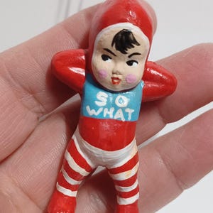May include: A vintage-style ornament of a baby in a red onesie with white stripes and a matching cap. The baby has a blue bib with the words "SO WHAT" in white. The ornament has a metal loop for hanging.
