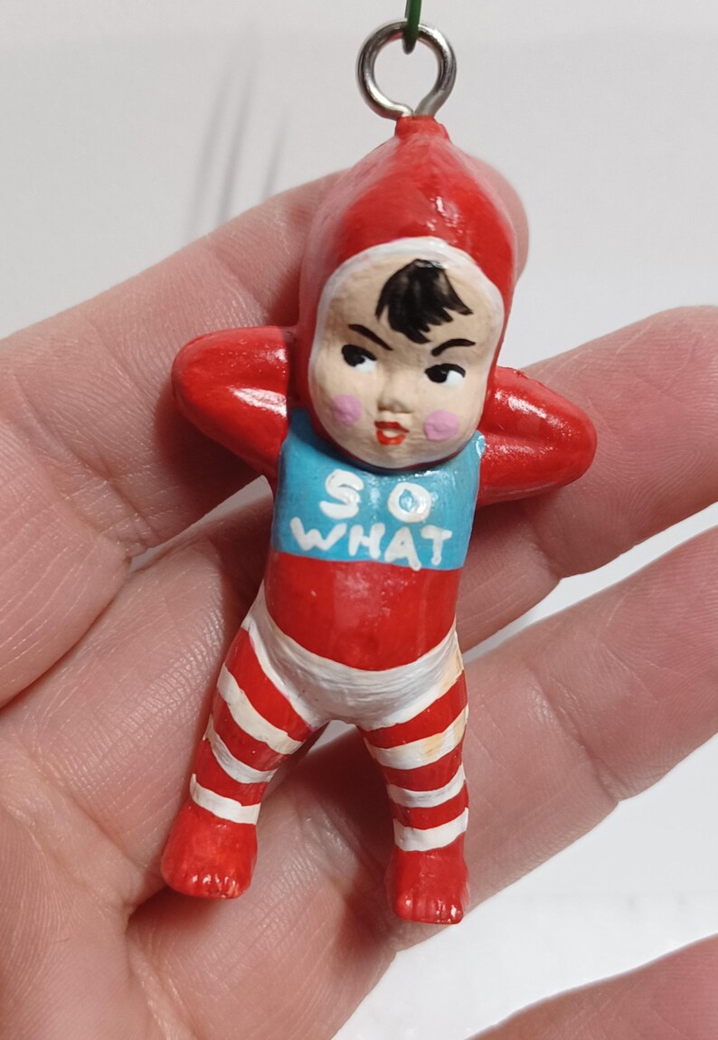 May include: A vintage-style ornament of a baby in a red onesie with white stripes and a matching cap. The baby has a blue bib with the words "SO WHAT" in white. The ornament has a metal loop for hanging.