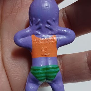 May include: A purple figurine of a person in a swimsuit. The figurine has an orange top and green and white striped bottoms. The text "MADE IN HONGKONG" is visible on the back of the figurine.