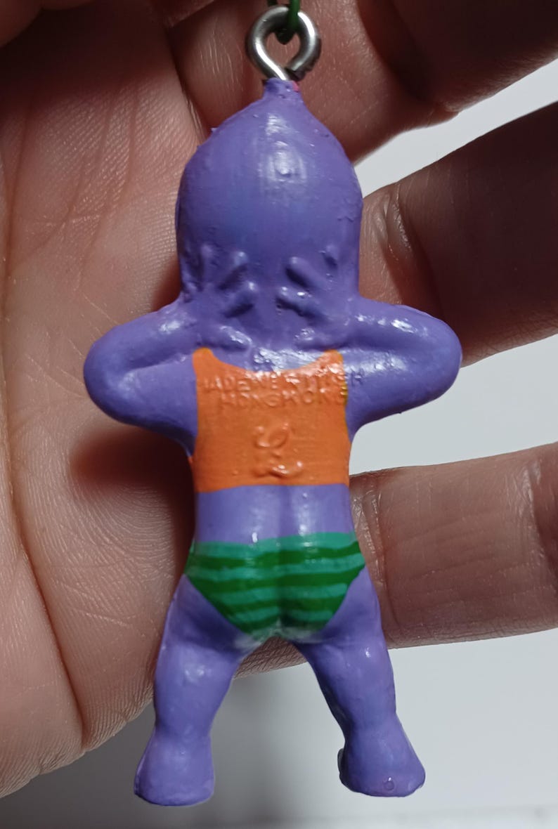 May include: A purple figurine of a person in a swimsuit. The figurine has an orange top and green and white striped bottoms. The text "MADE IN HONGKONG" is visible on the back of the figurine.