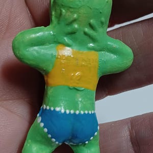May include: A green figurine of a person with arms raised, wearing a yellow top and blue shorts with white polka dots. The figurine has a small metal loop at the top for hanging.