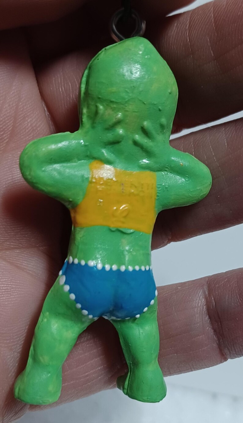 May include: A green figurine of a person with arms raised, wearing a yellow top and blue shorts with white polka dots. The figurine has a small metal loop at the top for hanging.