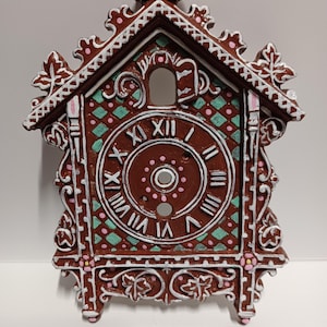 May include: A decorative brown clock with white and pink accents. The clock face features Roman numerals and a central hole. The clock has a gingerbread house design with a roof and decorative trim. The clock is a unique home decor item.