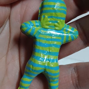 May include: A small, colorful figurine of a humanoid figure with arms crossed. The figure is painted with horizontal stripes of blue and green. The head, hands, and feet are solid green. A small metal ring is attached to the top.