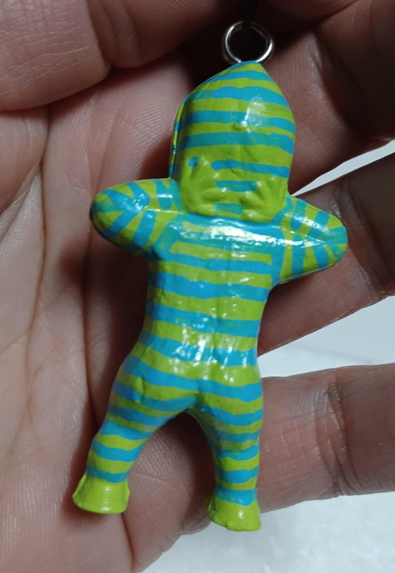 May include: A small, colorful figurine of a humanoid figure with arms crossed. The figure is painted with horizontal stripes of blue and green. The head, hands, and feet are solid green. A small metal ring is attached to the top.