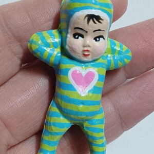 May include: A small, painted figurine of a baby in a striped blue and green onesie with a matching cap. The baby has a pink heart on its chest and is holding its arms up. The figurine has a metal loop at the top.