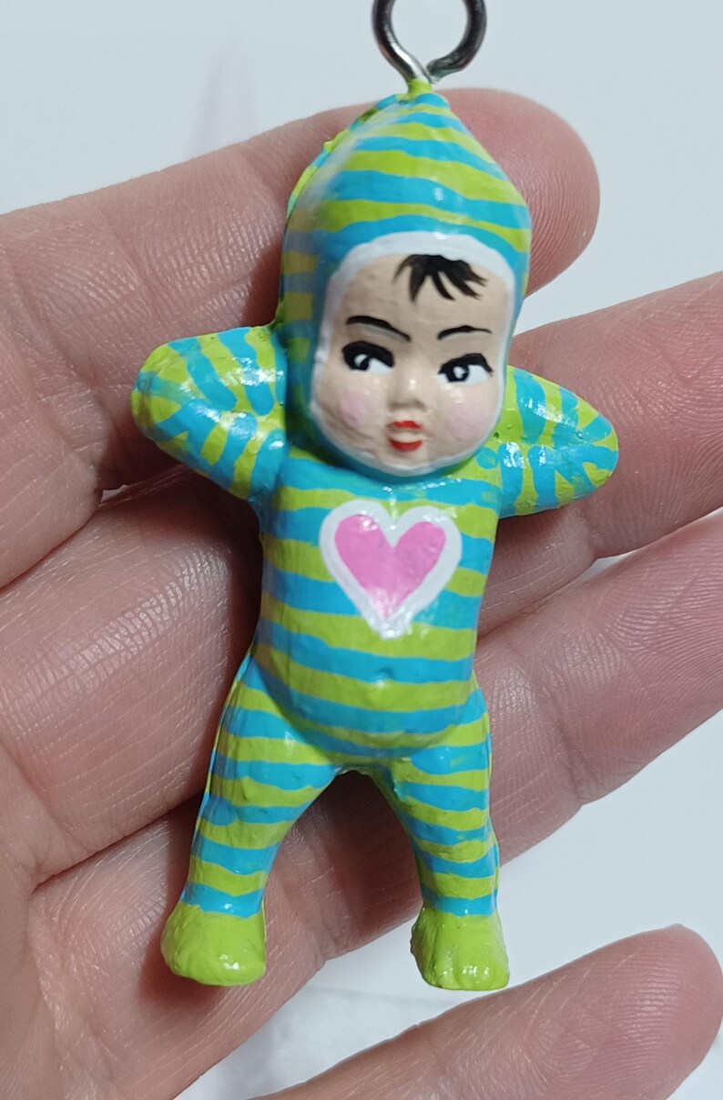 May include: A small, painted figurine of a baby in a striped blue and green onesie with a matching cap. The baby has a pink heart on its chest and is holding its arms up. The figurine has a metal loop at the top.