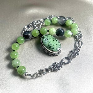 May include: A bracelet featuring a central oval-shaped green and black speckled stone pendant in a silver-toned setting, connected to a chain of round light green beads and dark grey beads, finished with a silver-toned metal link chain and hook clasp.