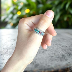 Puede incluir: Un anillo plateado con cinco piedras preciosas de colores: turquesa, turquesa, rojo y dos azules. El anillo se lleva en un dedo, mostrando los vibrantes colores de las piedras.