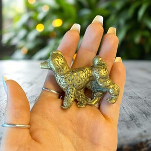 May include: A brass poodle figurine with detailed fur texture. The dog is in a standing pose, with its head and tail raised. The figurine is held in a hand, with rings on the fingers. The background is blurred with green and yellow bokeh.