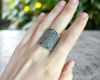 Puede incluir: Un anillo ancho, plateado, con una superficie texturizada, tachonado con pequeñas piedras oscuras. El anillo tiene forma rectangular y se lleva en una mano. Los bordes del anillo están bordeados con detalles triangulares.