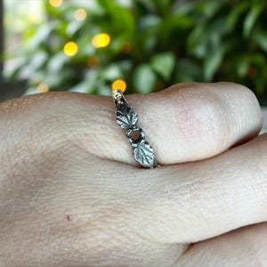 Puede incluir: Un anillo plateado con diseño de hojas. El anillo presenta tres hojas y una pequeña piedra redonda y marrón. Las hojas están detalladas con vetas grabadas. El anillo se muestra en el dedo de una persona.