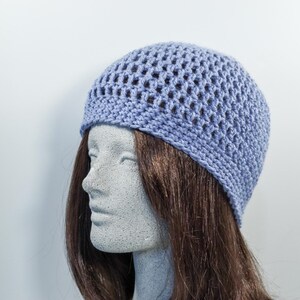May include: A periwinkle blue crochet beanie hat with an open weave design. The hat is displayed on a mannequin head with brown hair, set against a plain white background. The hat's texture and color are prominent.
