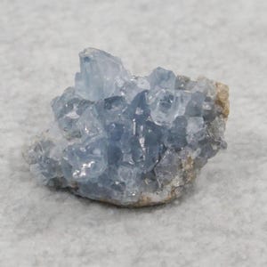 May include: A cluster of light blue celestite crystals. The crystals have a translucent appearance and are clustered together on a beige-colored matrix. The overall shape is irregular and natural, showcasing the mineral's crystalline structure.