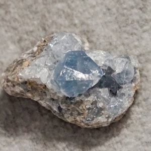 May include: A cluster of light blue celestite crystals embedded in a gray rock. The crystals have a sharp, geometric shape and a translucent appearance.
