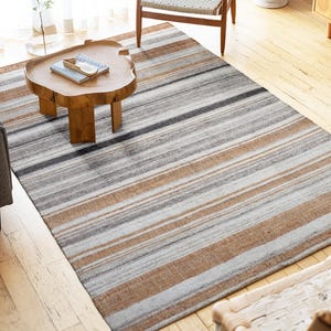 Pode incluir: Um tapete retangular com listras horizontais em tons de cinza, marrom e branco sujo. O design do tapete apresenta listras horizontais. O tapete é colocado sobre um piso de madeira. O tapete adiciona calor e textura ao ambiente.