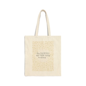 May include: Cream-colored tote bag with a floral pattern and the text "His mercies are new every morning." The bag has a rectangular shape with a handle for carrying. The design is simple and elegant.