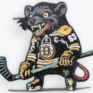 Puede incluir: Un parche negro con un dibujo de una rata de dibujos animados con una camiseta de hockey con el número 63. La rata sostiene un palo de hockey y tiene una expresión amenazante. La camiseta tiene detalles dorados y blancos y la letra "C".