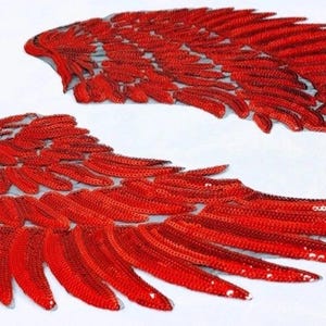Peut inclure: Deux appliques d'ailes rouges à paillettes, une complète et une partielle, sont présentées sur un fond blanc. Les ailes présentent un motif de plumes détaillé, avec des paillettes rouges et argentées.