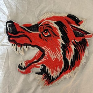 May include: An embroidered patch featuring the profile of a snarling wolf head. The design uses vibrant orange, black, and white thread to depict the animal with an open mouth, sharp teeth, and detailed fur texture, finished with a white border.