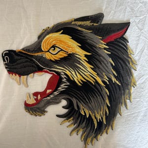 May include: Embroidered patch featuring a snarling wolf's head. The wolf has black, gray, and yellow fur, with red accents in the ear and mouth. The patch is detailed with sharp teeth and an open mouth.