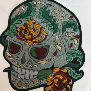 Könnte beinhalten: Ein großer gestickter Aufnäher mit einem grauen Totenkopf-Design, verziert mit grünen Blattmustern, gelben und roten Chrysanthemen sowie zwei roten rautenförmigen Akzenten in den Augenhöhlen, alles umrandet von einem dicken schwarzen Rand.