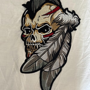 May include: Embroidered patch featuring a skull with a Native American headdress design. The skull is tan and black with red accents. The feathers are gray and white, outlined in black. The patch is likely for jackets or bags.
