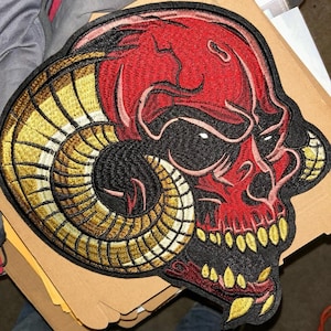 May include: Embroidered patch depicting a red skull with large, curved horns in gold and tan. The skull has a menacing expression with yellow teeth and is outlined in black.