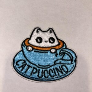 May include: Embroidered patch featuring a white cat in a blue teacup with the text "CATPUCCINO". The cat has large, round eyes and is surrounded by an orange rim. The teacup and saucer are light blue.