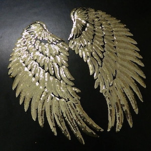 Peut inclure: Paire d'ailes d'ange dorées à paillettes sur fond noir. Les ailes sont détaillées avec des formes de plumes individuelles, créant un effet scintillant. Idéal pour les costumes ou la décoration.