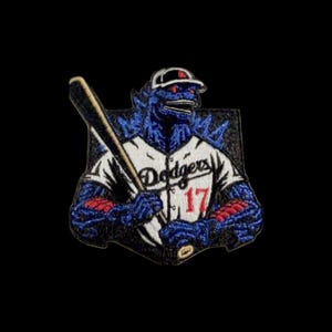 Puede incluir: Parche bordado con un monstruo azul con uniforme de béisbol de los Dodgers y gorra, sosteniendo un bate. La camiseta muestra "Dodgers" y el número 17 en rojo. El parche tiene un borde negro.