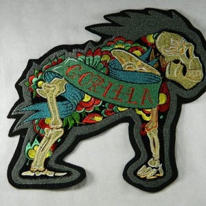 Puede incluir: Parche bordado con un diseño de esqueleto de gorila. El parche tiene un contorno negro y un borde gris. El esqueleto es dorado, con una pancarta que dice "GORILLA" en rojo. El fondo es una mezcla de patrones florales y de hojas coloridas.