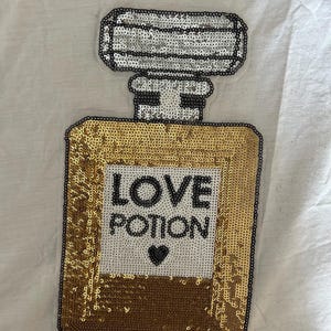 May include: A sequined patch in the shape of a perfume bottle. The bottle is gold with the words "LOVE POTION" and a black heart. The top of the bottle is silver.