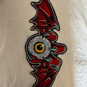 Puede incluir: Parche bordado con un ojo estilizado con iris amarillo, enmarcado por alas de murciélago rojas. El parche tiene un borde negro y está diseñado para su aplicación en ropa o accesorios. El diseño está detallado con varios colores y texturas.