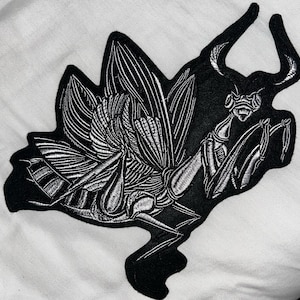 May include: An embroidered patch featuring a detailed praying mantis design. The insect is rendered in white thread against a solid black fabric background, showcasing intricate line work on the wings, segmented body, and curved antennae.