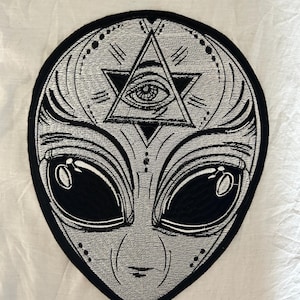 May include: Embroidered patch featuring an alien face with large black eyes and a triangular symbol with an eye at the top. The patch is primarily silver and black, with intricate detailing and a black border. The design has a mystical and otherworldly theme.