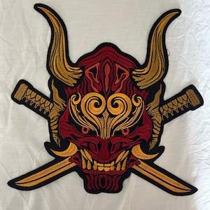 Könnte beinhalten: Gestickter Aufnäher mit einer rot-schwarzen Oni-Maske mit goldenen Hörnern und Details, gekreuzt von zwei goldenen Katanas. Der Aufnäher hat ein detailliertes Design und eignet sich für Jacken, Taschen oder andere Kleidungsstücke.
