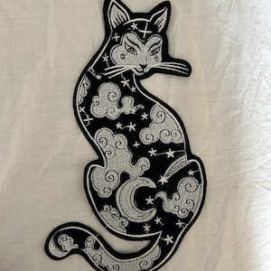 May include: Black and white embroidered cat patch with celestial design. The cat features a crescent moon, stars, and cloud patterns. The patch has a detailed outline and is likely for clothing or accessories.