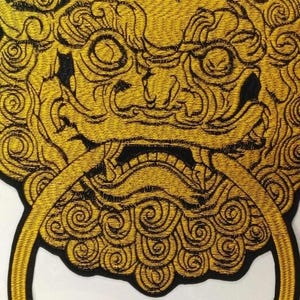 May include: Embroidered patch featuring a golden lion-dog design with a circular handle. The patch has a black border and intricate details, suitable for jackets, bags, or other fabric items.