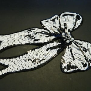 May include: A decorative bow appliqué featuring white sequins with black sequin trim and accents. The centre of the bow is embellished with small white faux pearls and silver beads, creating a textured, high-contrast design for garment embellishment.