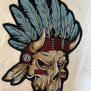 May include: Embroidered patch featuring a skull wearing a Native American headdress. The skull is tan and cream-colored, with red and blue accents. The headdress has blue feathers and a red band. The patch is outlined in black.