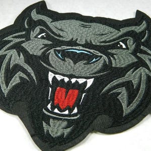May include: Embroidered patch featuring a snarling wolf head. The patch is primarily black with gray and white accents, including the wolf's fur, teeth, and eyes. The wolf's mouth is open, revealing a red tongue.