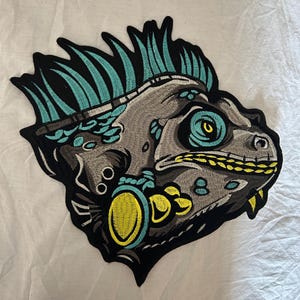 Op de afbeelding: Geborduurde patch met een gestileerde hagedissenkop met gedetailleerde kenmerken. De patch heeft een zwarte rand, grijze en blauwgroene accenten en gele highlights. Het oog van de hagedis is helder turquoise.